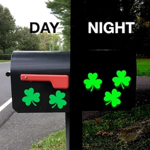 May include: A black mailbox with a red handle and four green shamrock stickers. The mailbox is shown in both daylight and nighttime, with the shamrocks glowing in the dark.