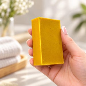 May include: A rectangular bar of solid soap with a vibrant mustard yellow color and a smooth, matte texture. The soap features subtle, fine-grained flecks throughout its surface, indicating a natural or herbal composition in its appearance.