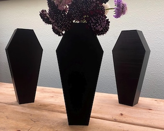 May include: Three black coffin-shaped vases on a wooden surface. The vase in the center holds a bouquet of purple, white, and pink flowers.