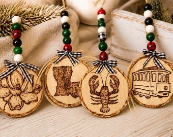 May include: Four round wooden ornaments featuring wood-burned designs of a magnolia flower, the state of Louisiana, a lobster, and a streetcar. Each ornament is topped with a black and white gingham ribbon and a string of red, green, black, and white beads.