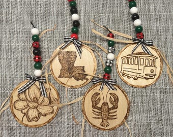 May include: Four wooden ornaments with black and white checkered ribbon and red and green beads. The ornaments feature a magnolia flower, a lobster, the state of Louisiana, and a streetcar.