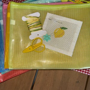 May include: A stack of translucent mesh zipper pouches in various colors including yellow, pink, and blue. On top sits a white spool of variegated yellow and green embroidery thread, a small yellow pair of scissors, and a square of white canvas.