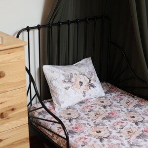 May include: A black metal bed frame with a white floral patterned sheet and pillow. The bed is made up with a white pillow with a floral design. The bed is in a room with a wooden dresser and a green curtain.