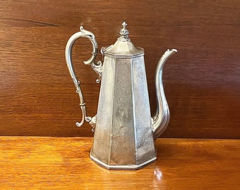 Op de afbeelding: Een zilveren theepot met een achthoekig lichaam en een decoratief handvat. De theepot heeft een gedetailleerd gegraveerd ontwerp en een knop op de deksel.