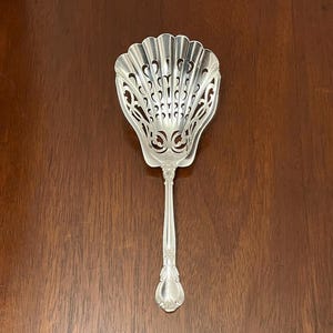 Puede incluir: Una cuchara de servir plateada con un cuenco decorativo en forma de abanico y un mango largo y ornamentado. El cuenco presenta un diseño calado y el mango tiene un detallado patrón floral. Este utensilio vintage es probablemente para servir azúcar u otros productos secos.