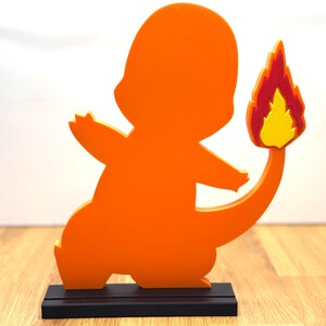 Op de afbeelding: Een oranje Charmander-figuur, een populair personage uit de Pokémon-franchise, wordt weergegeven op een zwarte rechthoekige basis. De figuur heeft een vlam aan het einde van zijn staart, met gele, rode en oranje kleuren. De figuur is gemaakt van een solide materiaal.