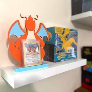 May include: An orange and light blue Dragonite-shaped display stand holding a Dragonite trading card in a clear protective case. Next to it sits a blue and yellow box labeled Pokemon Trading Card Game Ascended Heroes on a white shelf.