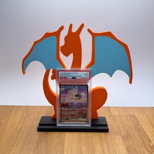 May include: A PSA graded 2023 Pokemon Mew En Charizard ex #199 Mint 9 trading card in a clear plastic slab, displayed on a black stand in front of an orange and light blue dragon-shaped cutout silhouette with a long tail and large wings.