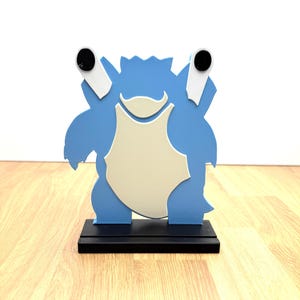 May include: A blue and cream-colored Blastoise figure, a popular character from the Pokémon franchise, stands on a black rectangular base. The figure has a light blue body with a cream-colored chest and two black circular details.