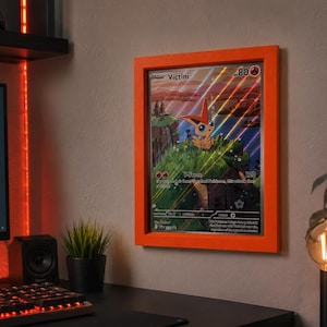 May include: A rectangular orange frame containing a large Victini trading card. The card features a holographic finish with diagonal light streaks, displaying the character Victini, the text Victini, 80 HP, and V-Force 120, mounted on a wall.