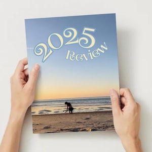 Op de afbeelding: Een tijdschriftomslag met de tekst "2025 Review" in een lichtblauwe letter. De afbeelding op de omslag toont een strandscène bij zonsondergang, met een persoon op de kust. Het tijdschrift wordt door twee handen vastgehouden.