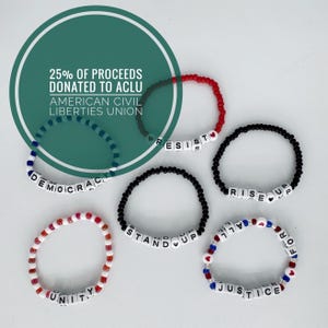May include: A collection of beaded bracelets in red, white, black, and blue. The bracelets spell out words like "DEMOCRACY", "RESIST", "UNITY", "STAND UP", "RISE UP", and "JUSTICE FOR ALL". The image also includes text that says "25% OF PROCEEDS DONATED TO ACLU".