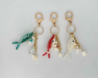 May include: Three keychains featuring beaded fish charms in green, red, and white. Each fish is attached to a gold-tone lobster clasp and ring, with additional small dangling beads and shell-like accents hanging from the gold metal chain links.