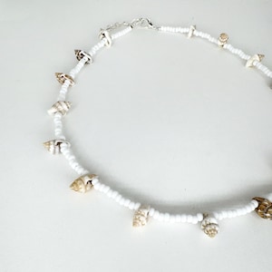May include: White beaded necklace with small seashells. The necklace is made of white beads and has small seashells attached to it. The seashells are a light brown color.