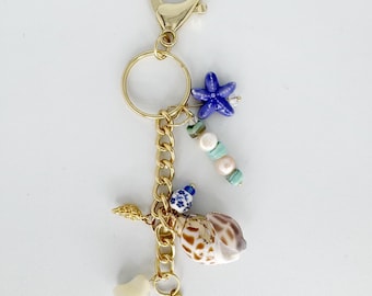 May include: Gold-tone keychain with a variety of beach-themed charms. Includes a blue starfish, seashells, pearls, and a small gold starfish charm. The keychain is attached to a gold clasp.