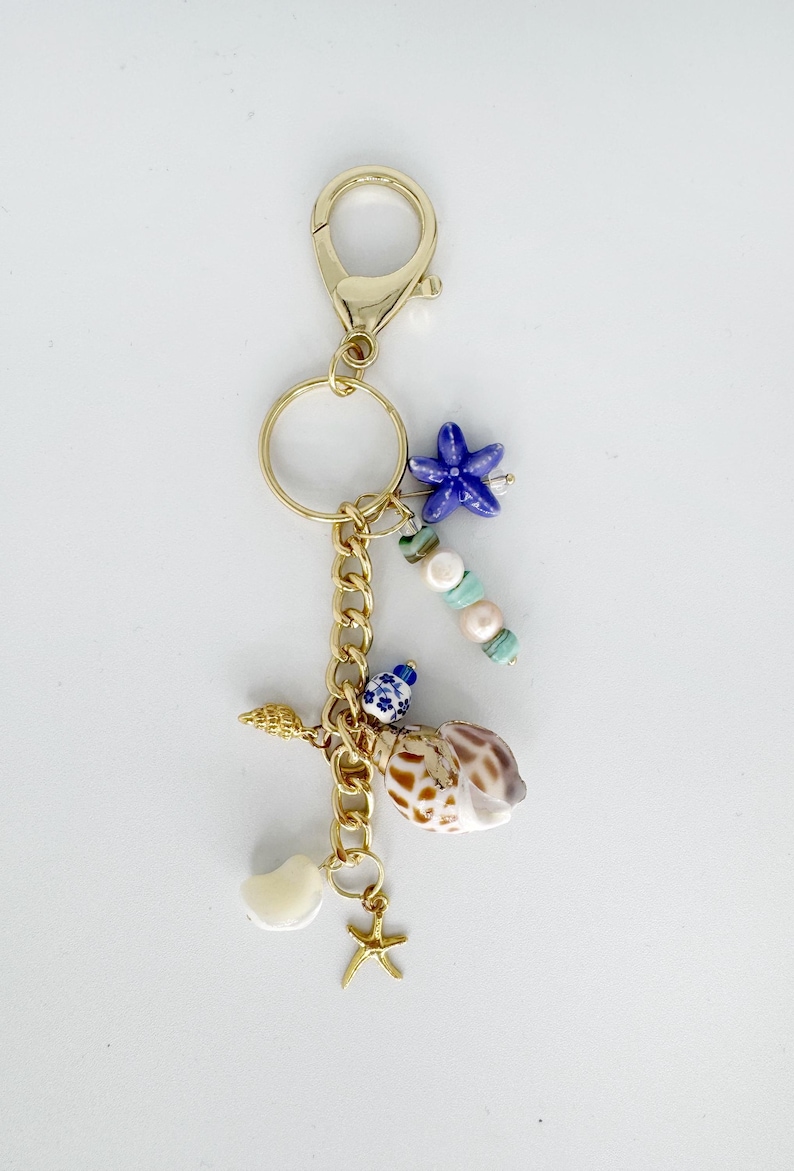 May include: Gold-tone keychain with a variety of beach-themed charms. Includes a blue starfish, seashells, pearls, and a small gold starfish charm. The keychain is attached to a gold clasp.