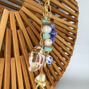 May include: A bamboo handbag with a gold handle and a decorative charm. The charm features a seashell, a starfish, and various beads in shades of blue, green, and pearl. The seashell has gold accents.