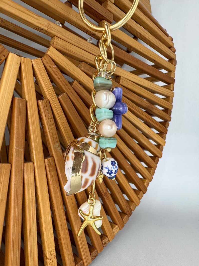 May include: A bamboo handbag with a gold handle and a decorative charm. The charm features a seashell, a starfish, and various beads in shades of blue, green, and pearl. The seashell has gold accents.