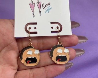 May include: A pair of handmade earrings featuring a cartoon character with a surprised expression. The earrings have gold-colored hooks and are attached to a white card with the words "Handmade Earrings" and a signature. The character's face is tan with black hair and a wide open mouth.