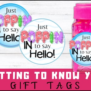 Puede incluir: Dos etiquetas redondas para regalos con el texto "Just Poppin' In to say Hello!" en letras de colores sobre un fondo azul. Las etiquetas están unidas a una botella rosa de solución de burbujas.
