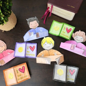 May include: Five paper craft figures of children holding open books with the words "I love the Scriptures" on the cover. The books are in different colors, including green, orange, pink, and blue. The figures are all smiling and have different hair colors.