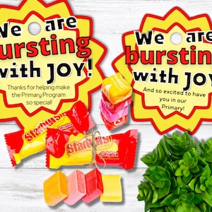 May include: Two yellow star-shaped tags with red text that reads "We are bursting with JOY!" One tag says "Thanks for helping make the Primary Program so special!" and the other says "And so excited to have you in our Primary!" The tags are on a wood background with candy and a small green plant.