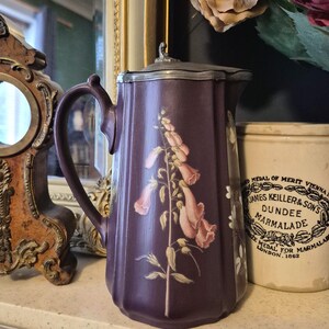 May include: A vintage purple pitcher with a silver lid and handle. The pitcher is decorated with a floral design in shades of pink and white. A white jar with text is visible in the background.
