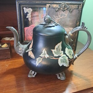 May include: Black teapot with silver accents and painted ivy leaves and a butterfly. The teapot has three feet and a knob on the lid.