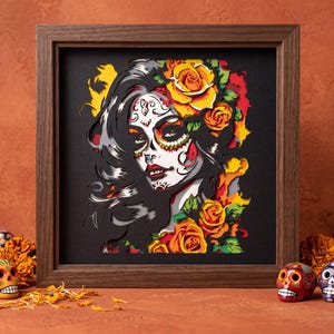 May include: A multi-layered paper art piece in a dark wood frame depicting a person with sugar skull face paint, dark hair, and yellow and orange roses. The artwork features intricate cutouts in black, white, red, yellow, and green layers.