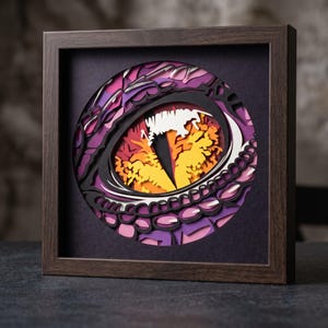 May include: A square dark wood frame containing a multi-layered paper art piece. The artwork features a circular design of a reptilian eye with a vertical pupil, rendered in shades of purple, pink, orange, and yellow with black outlines.