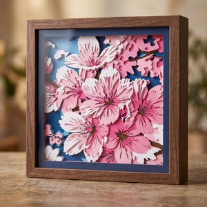 May include: A square wooden shadow box frame containing a multi-layered paper art piece. The artwork features intricate, laser-cut cherry blossom flowers in various shades of pink and white, set against a deep blue background.