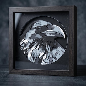 May include: A layered paper art piece featuring a raven head in profile, encased in a dark brown square wooden frame. The artwork uses shades of black, gray, and white to create depth, set against a circular black background within the frame.
