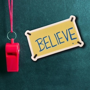 Peut inclure: Un sifflet rouge avec un cordon suspendu à un tableau vert avec une pancarte jaune sur laquelle est inscrit "BELIEVE" en lettres bleues.