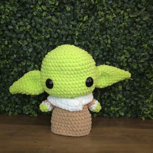 Pode incluir: Boneca de croch&ecirc; feita &agrave; m&atilde;o, semelhante a um personagem popular. A boneca apresenta uma cabe&ccedil;a verde clara e orelhas grandes, olhos pretos, uma gola branca e um corpo bege. A boneca &eacute; exibida em um fundo verde.