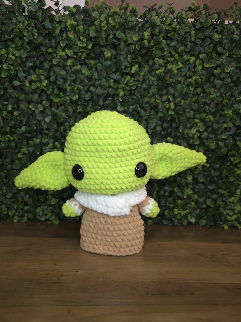 Pode incluir: Boneca de croch&ecirc; feita &agrave; m&atilde;o, semelhante a um personagem popular. A boneca apresenta uma cabe&ccedil;a verde clara e orelhas grandes, olhos pretos, uma gola branca e um corpo bege. A boneca &eacute; exibida em um fundo verde.