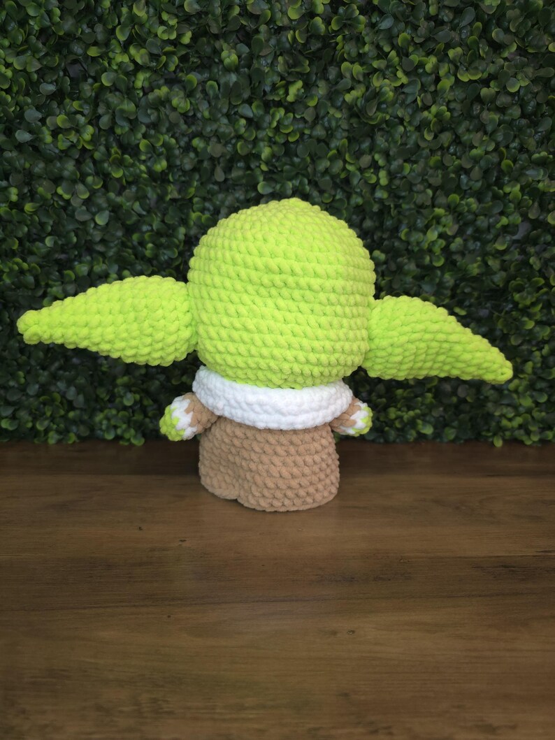 Pode incluir: Boneca de croch&ecirc; feita &agrave; m&atilde;o de um personagem com grandes orelhas verdes, uma cabe&ccedil;a verde e um corpo marrom. A boneca tem uma gola branca e m&atilde;os e p&eacute;s pequenos. A boneca est&aacute; sentada em uma superf&iacute;cie de madeira com um fundo verde frondoso.