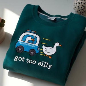 May include: Teal sweatshirt with a cartoon duck design. A blue police car with a duck inside is chasing another duck. The text "got too silly" is below the graphic. The sweatshirt is folded on a white surface.