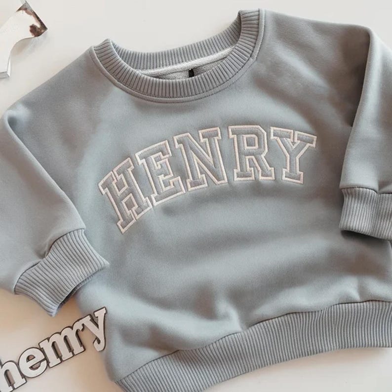 Puede incluir: Sudadera azul claro con el nombre "HENRY" bordado en letras blancas en el pecho. La sudadera tiene pu&ntilde;os y dobladillo de canal&eacute;. La palabra "henry" tambi&eacute;n es visible en una pieza separada.