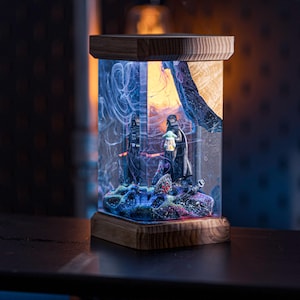 Puede incluir: Una base de l&aacute;mpara de madera con un cilindro de resina transparente que contiene una escena con tem&aacute;tica de Star Wars. La escena presenta a Darth Vader y Grogu, con un fondo de galaxia.