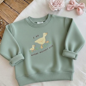May include: Pale green sweatshirt featuring a duck and duckling graphic with the text "I GO WHERE MOMMY GOES". The sweatshirt has a crew neck and ribbed cuffs and hem.