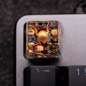 Pode incluir: Uma tecla de teclado personalizada com um design detalhado de capacete dourado e laranja. A tecla tem um acabamento transparente e texturizado, sobre um fundo escuro de teclado. O design é intrincado e atraente.