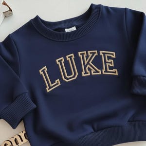 Puede incluir: Sudadera azul marino con el nombre "LUKE" bordado en letras doradas de estilo universitario. La sudadera tiene cuello redondo y pu&ntilde;os y dobladillo acanalados. La tela parece suave y c&oacute;moda.