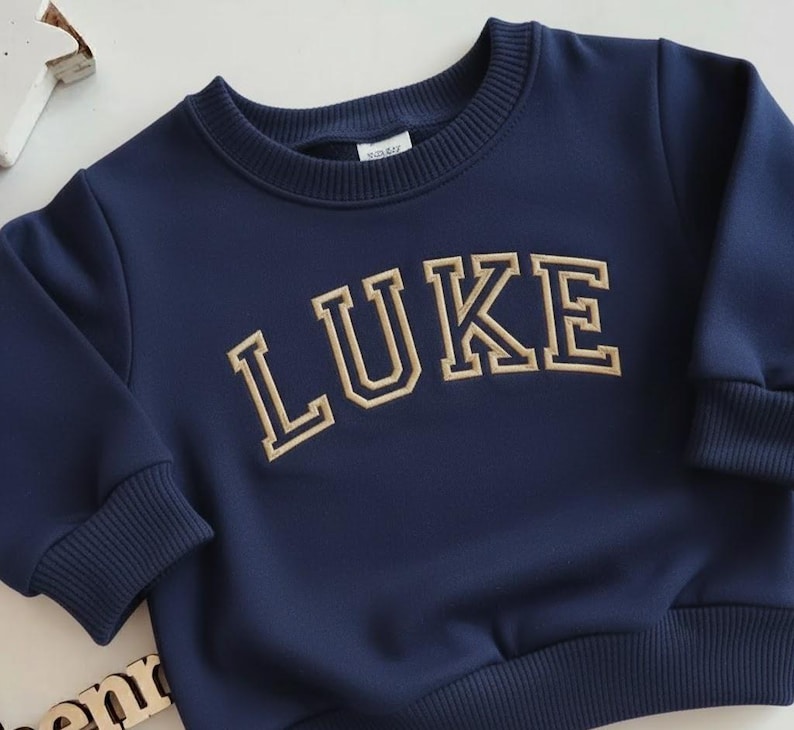 Puede incluir: Sudadera azul marino con el nombre "LUKE" bordado en letras doradas de estilo universitario. La sudadera tiene cuello redondo y pu&ntilde;os y dobladillo acanalados. La tela parece suave y c&oacute;moda.