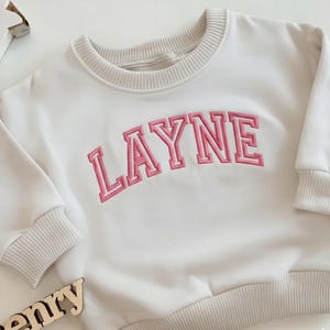 Puede incluir: Sudadera color crema con el nombre "LAYNE" bordado en letras rosas. La sudadera tiene cuello redondo, pu&ntilde;os y dobladillo de canal&eacute;. Las mangas son largas y la tela parece suave.