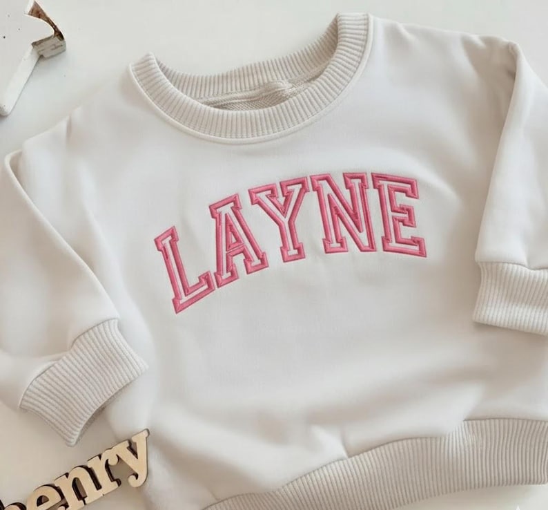Puede incluir: Sudadera color crema con el nombre "LAYNE" bordado en letras rosas. La sudadera tiene cuello redondo, pu&ntilde;os y dobladillo de canal&eacute;. Las mangas son largas y la tela parece suave.