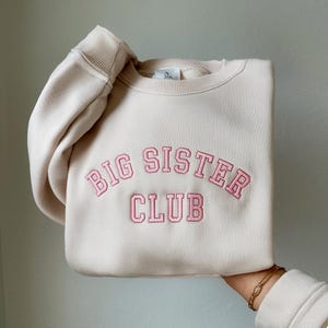 K&ouml;nnte beinhalten: Ein cremefarbener Pullover mit dem gestickten Schriftzug "BIG SISTER CLUB" in Rosa.