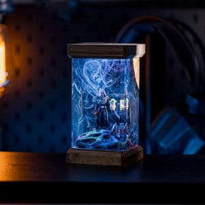 Puede incluir: Una l&aacute;mpara de resina azul oscuro con base de madera presenta una figura de Star Wars de Darth Vader de pie en una resina azul y blanca en espiral. La l&aacute;mpara est&aacute; iluminada desde el interior, proyectando un brillo azul.