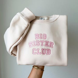 K&ouml;nnte beinhalten: Ein cremefarbener Pullover mit dem gestickten Text "BIG SISTER CLUB" in Rosa.