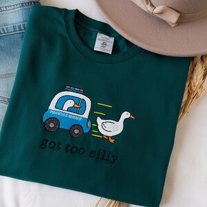 May include: Teal green t-shirt with an embroidered design of a cartoon goose in a police car chasing another goose. The text "got too silly" is below the design. A pair of blue jeans and a beige hat are also in the image.