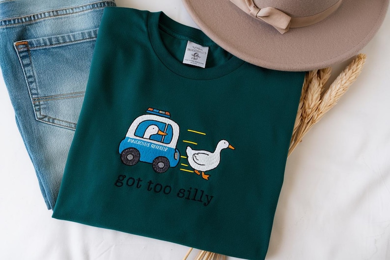 May include: Teal green t-shirt with an embroidered design of a cartoon goose in a police car chasing another goose. The text "got too silly" is below the design. A pair of blue jeans and a beige hat are also in the image.